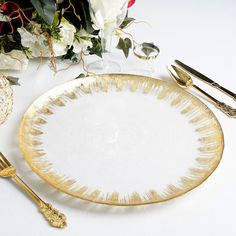 there is a white and gold plate next to some silverware on the table with flowers in the background