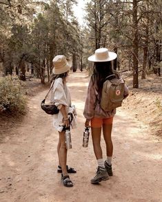 two young women walking down a dirt road in the woods with backpacks and hats on