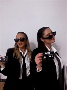 two women in suits and sunglasses holding up small camera's with their eyes closed