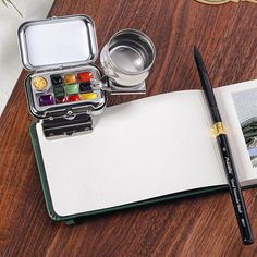 an open notebook with watercolors and a pen on it sitting on a table