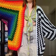 a woman standing in front of a crocheted blanket holding up a rainbow afghan