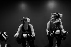 two women on exercise bikes in a black and white photo, one smiling at the camera