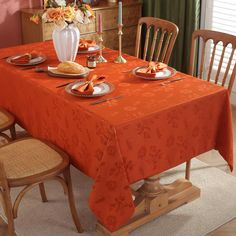 a dining room table with an orange table cloth and place settings on it, in front of a window