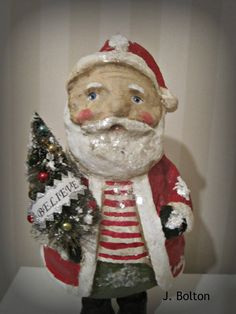 a santa clause figurine holding a small christmas tree in his right hand, with the words believe written on it