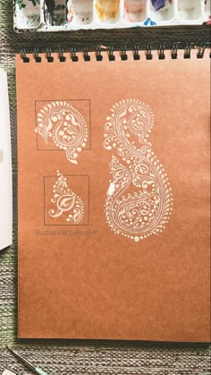 a brown notebook with white paisley designs on it next to paintbrushes and watercolors