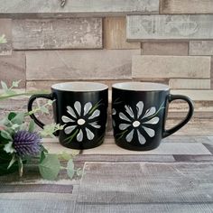 two black coffee mugs with white flowers on them sitting next to a purple flower