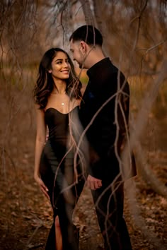 a man and woman standing next to each other in front of some trees with leaves on the ground