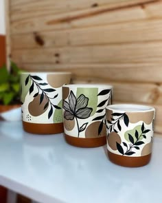 three coffee cups sitting on top of a white counter next to a potted plant