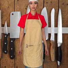 a woman wearing an apron and hat standing in front of knives on a wooden wall