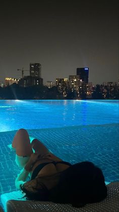 a person laying on the edge of a swimming pool at night with city lights in the background