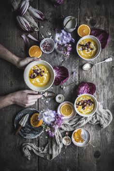 two hands holding bowls of soup with oranges, onions and other food items on the table