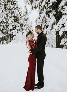 a man and woman standing in the snow with trees behind them, looking at each other