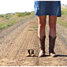 a woman standing on top of a dirt road next to a pair of cowboy boots