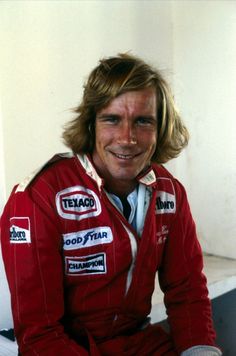 a man with long hair sitting on the ground wearing a red racing suit and smiling