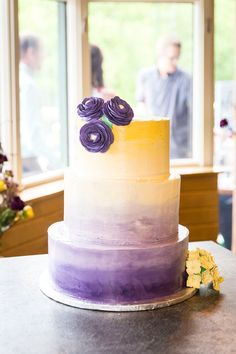 a multi - layered cake with purple flowers on top sits on a table in front of a window