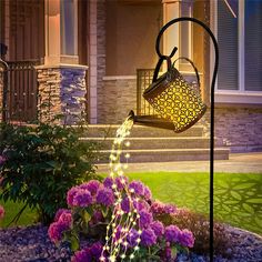 a watering can is lit up with fairy lights in front of some flowers and bushes