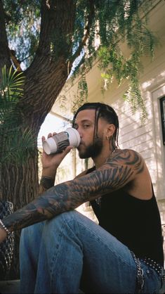 a man sitting under a tree drinking from a water bottle with tattoos on his arm