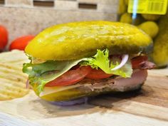 a close up of a sandwich on a cutting board with pickles and tomatoes in the background