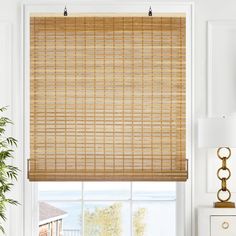 a window with bamboo blinds in a living room next to a white dresser and potted plant