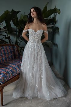 a woman standing in front of a couch wearing a wedding dress with an off the shoulder design