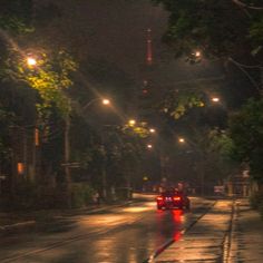 a red car driving down a wet street at night with lights shining on the trees