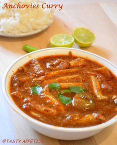 a white bowl filled with curry next to rice and limes on a wooden table