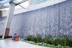 two people are sitting on a bench in front of a waterfall at the entrance to a building