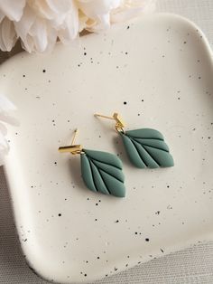 two green leaf shaped earrings sitting on top of a white plate next to a flower