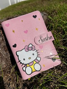 a pink hello kitty notebook sitting on top of grass next to a tree trunk with hearts