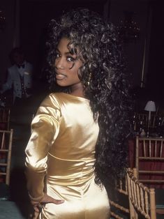 a woman with long black hair sitting in a chair wearing a gold dress and large hoop earrings