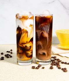 two glasses filled with ice and coffee on top of a counter next to some coffee beans