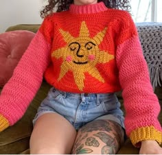 a woman sitting on top of a green couch wearing a red sweater with a yellow sun drawn on it