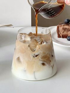 someone pouring coffee into a glass with ice and blueberries on the table next to a slice of cake