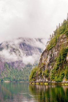 the mountains are covered in fog and clouds above water with trees on both sides of them