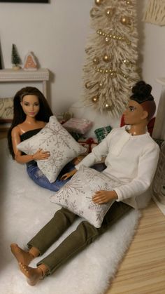 two dolls are sitting on the floor in front of a christmas tree, one is holding a pillow