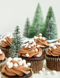 cupcakes with frosting and marshmallows are on a table next to small trees