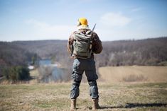 a man with a backpack and knife in his hands standing on the grass looking at mountains