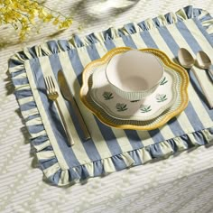 a place setting on a striped tablecloth with yellow and white dishes, silverware and flowers