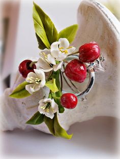 a ring with cherries and leaves on it sitting in a shell shaped vase next to a flower