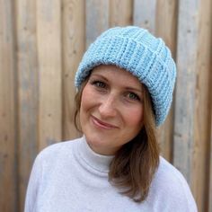 a woman wearing a blue knitted beanie smiles at the camera while standing in front of a wooden fence