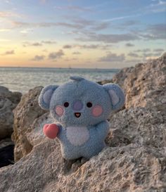 a small stuffed koala bear sitting on top of a rock near the ocean at sunset