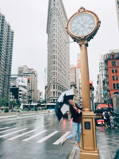 a woman holding an umbrella while standing next to a clock on a pole in the middle of a city