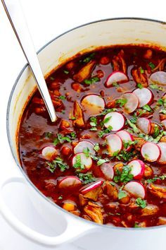 a white pot filled with stew and radishes on top of a table next to a spoon