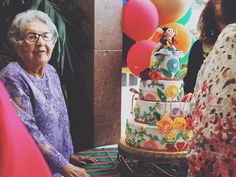 an older woman sitting in front of a cake on top of a table next to balloons