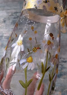 a hand holding a clear glass vase with flowers and bees painted on the side,