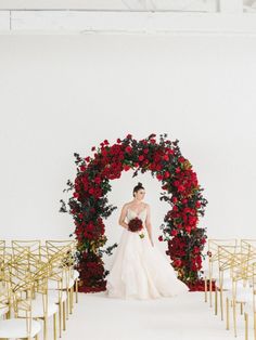 the bride is standing in front of an arch with red roses on it and gold chairs