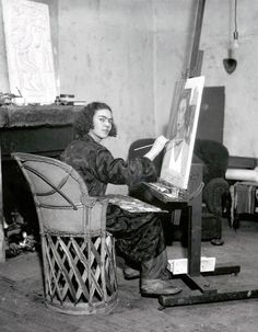 an old photo of a woman sitting in front of a easel with a painting on it