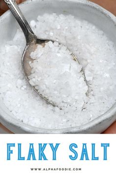 a spoon full of flaky salt in a white bowl with the words flaky salt on it