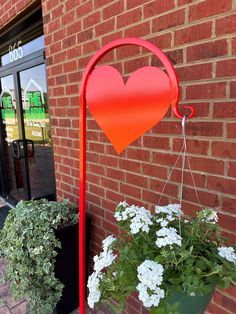 a red heart shaped sign hanging from the side of a brick building next to potted plants