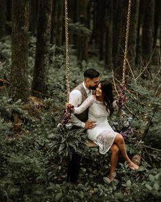 a man and woman are sitting on a swing in the woods, holding each other